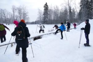 ryhmä naisia harjoittelee lumisessa maisemassa matalan puuaidan ylitystä, miesopettaja seuraa harjoitusta
