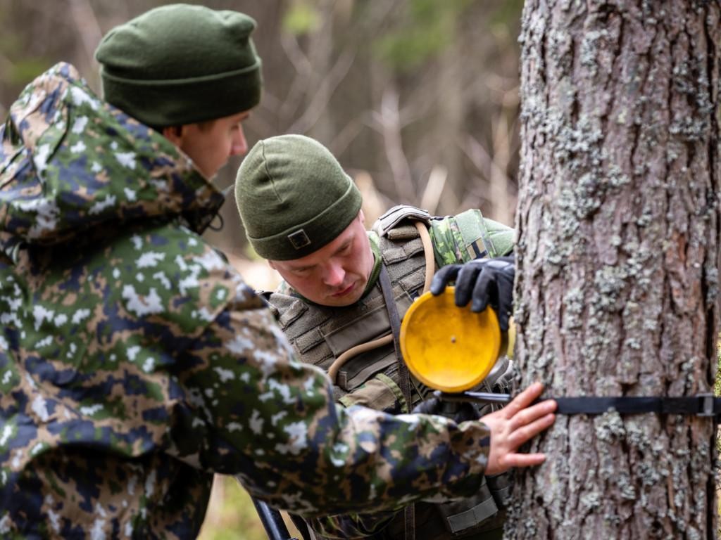 Kaksi miestä metsässä maastopuvuissa harjoittelevat viuhka- ja kylkimiinojen laittoa
