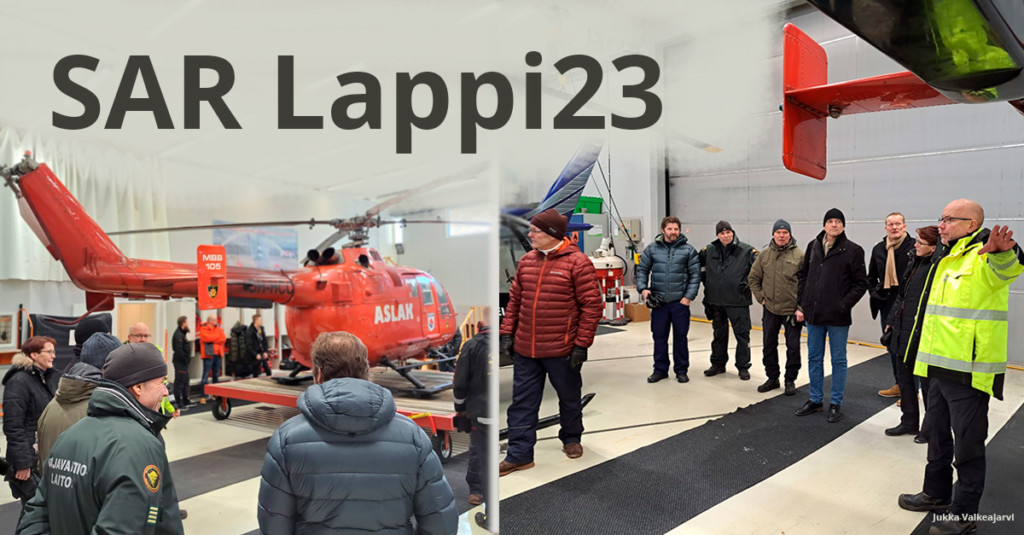 Kuvassa SAR Lappi 23 -harjoituksen valmistelijota kuuntelemassa esitelmää. Taustalla pelastushelikopteri.