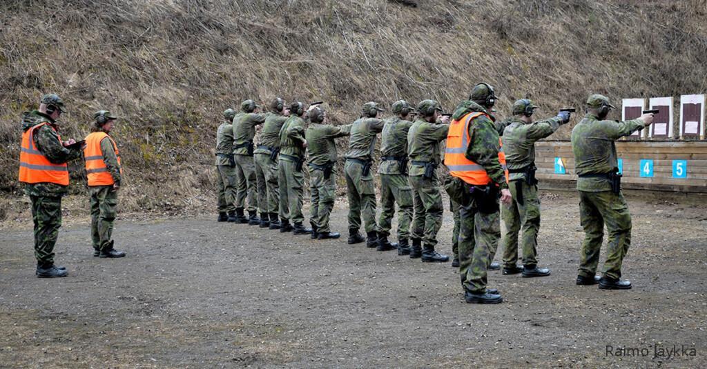 Kolme oransseissa liiveissä olevaa ammunnanjohtajaa seuraa rivissä olevien kurssilaisten pistooliampumista kohti maalitauluja.