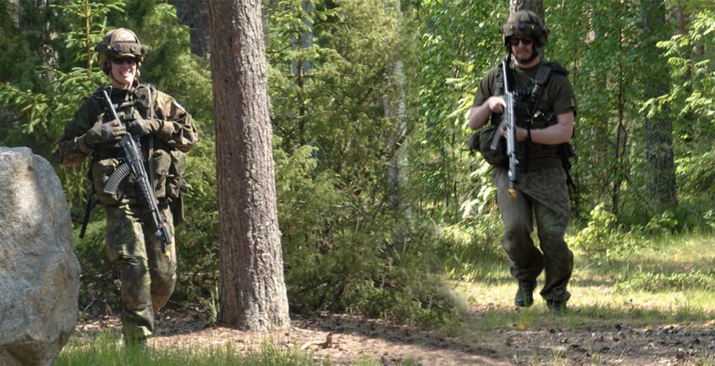 Kaksi taistelijaa juoksee metsässä aseet kädessä. Metsäsä kasvaa mantyjä, kuusia ja lehtipuita.