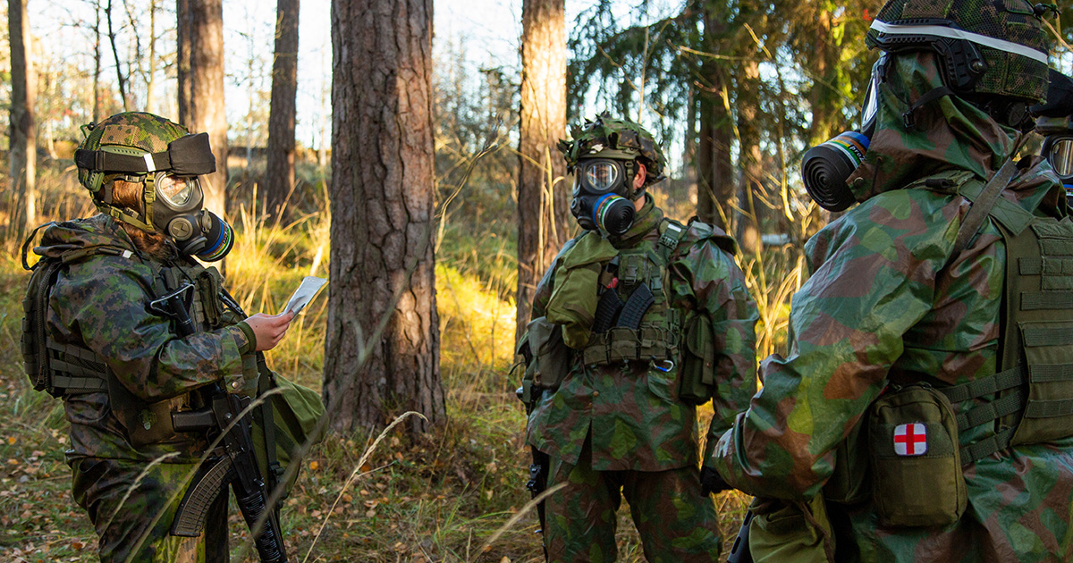 Kolme maastoasuista henkilöä suojanaamarit kasvoillaan keskustelevat metsässä.