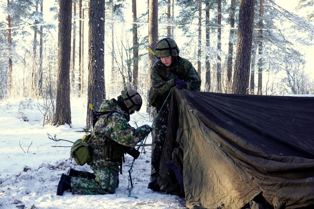 Kaksi naista maastopuvuissa pystyttää telttaa talvisessa metsässä.