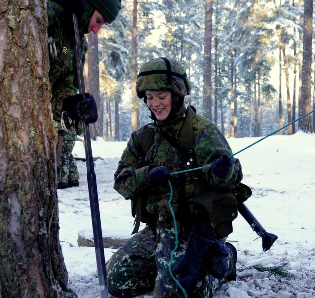 Nainen maastopuvussa ulkona pakkasessa kiinnittämässä telttanarua.