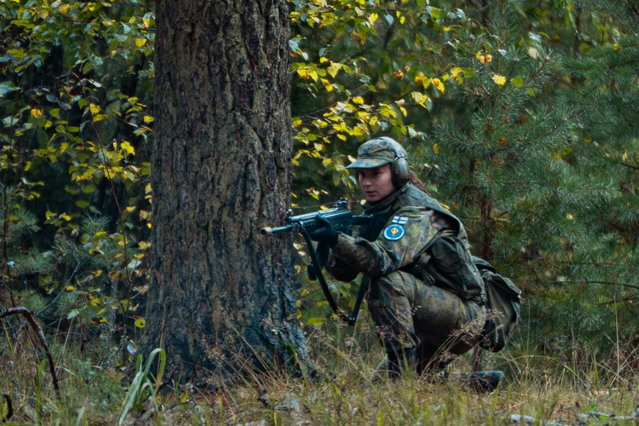 Maastopukuinen henkilö on kyykyssä puun juuressa ja tähtää aseellaan.