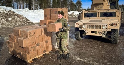 Sotilasasuinen nainen kantamassa pahvilaatikoita. Taustalla ulkomaalainen sotilasajoneuvo