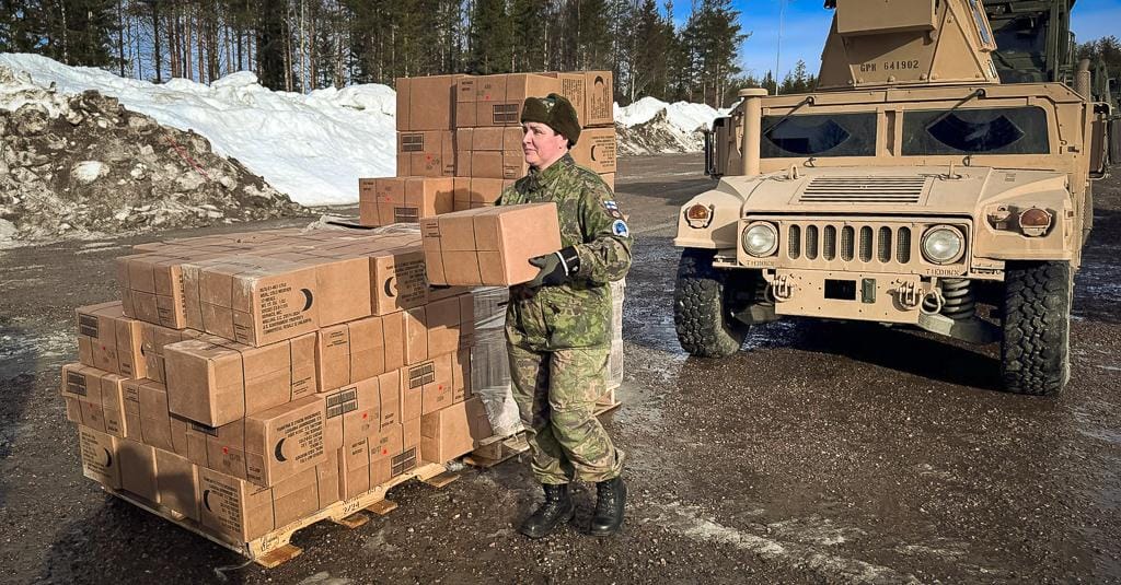 Sotilasasuinen nainen kantamassa pahvilaatikoita. Taustalla ulkomaalainen sotilasajoneuvo