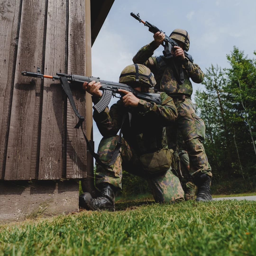 Kaksi maastopukuista henkilöä tähtäävät aseilla talon nurkalla.