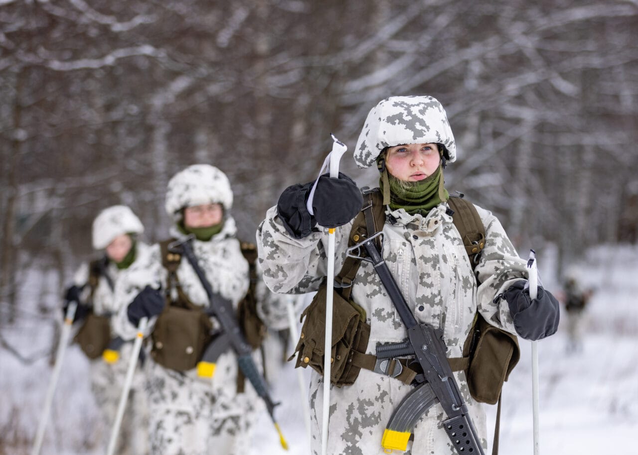 Kolme naista valkoisissa maastopuvuissa hiihtää lumisen maiseman keskellä.