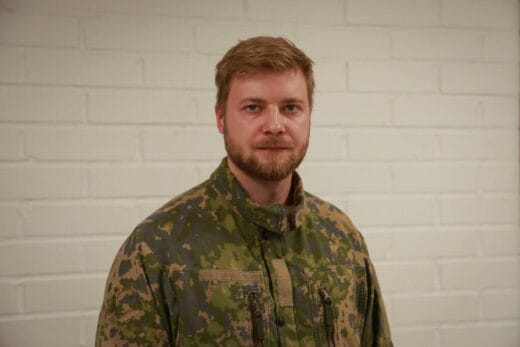Maastopukuinen mies seisoo valkoisen tiiliseinän edessä.