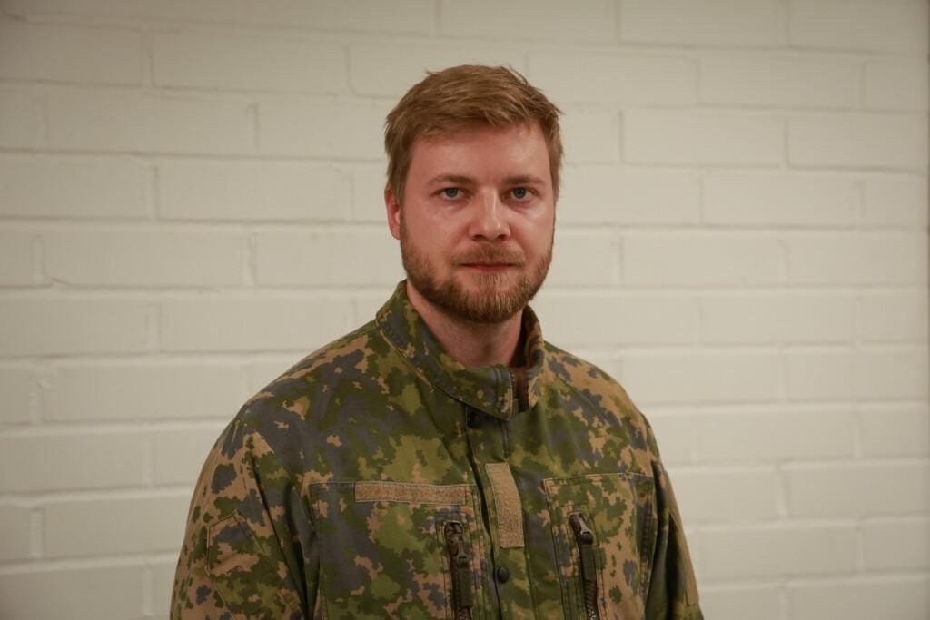 Maastopukuinen mies seisoo valkoisen tiiliseinän edessä.
