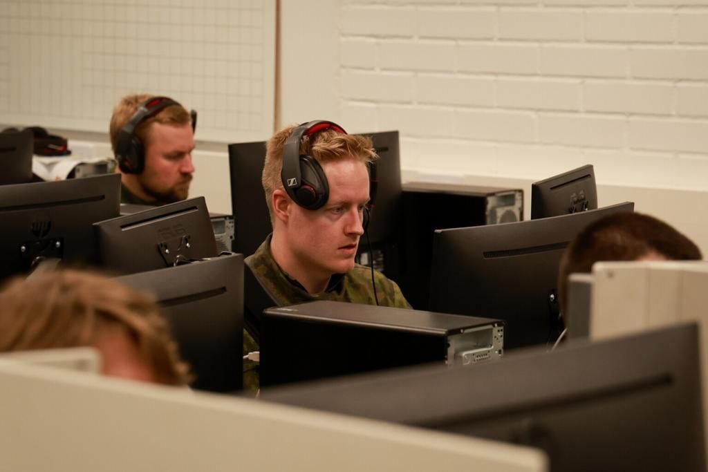 Maastopukuun pukeutunut mies istuu tietokoneen ääressä kuulokkeet päässä.