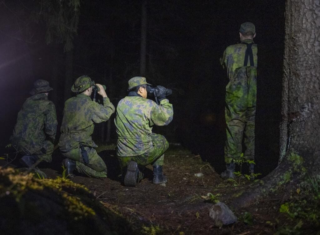 Neljä sotilasta tähystämässä pimeään metsään takaapäin kuvattuna.