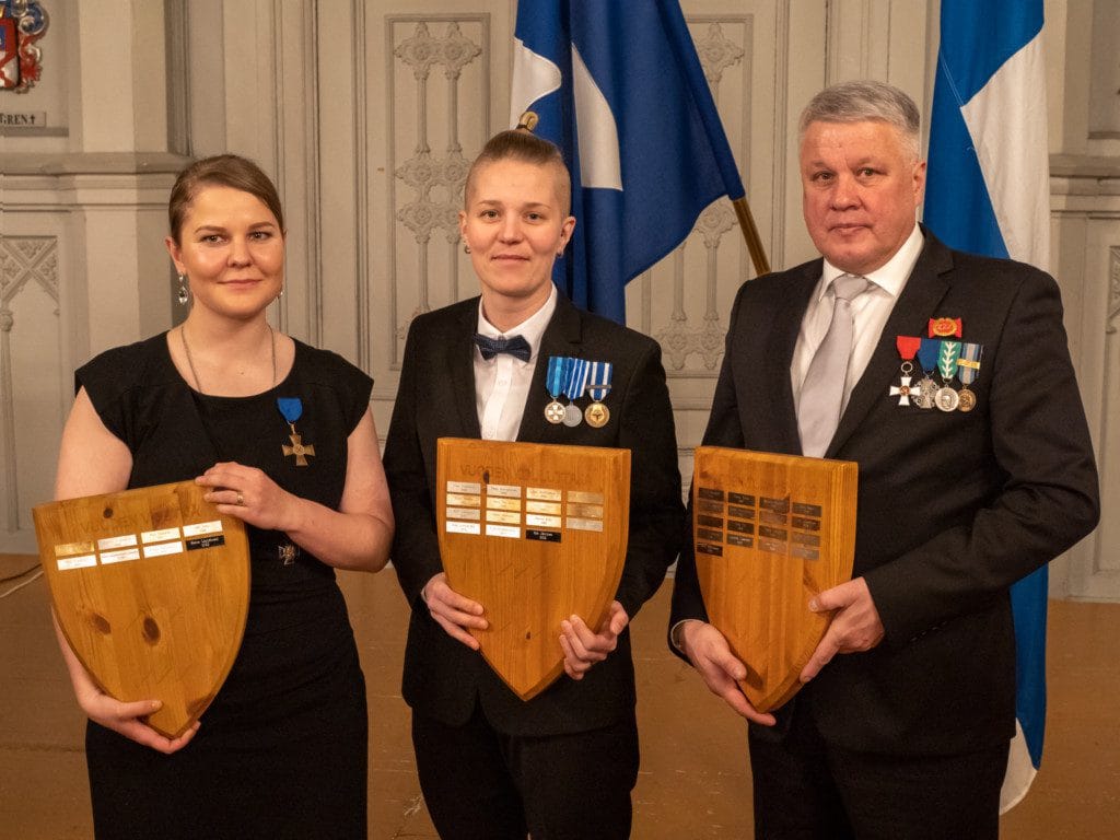 Vuoden viestijä, kouluttaja ja toimihenkilö seisovat käsissään kunniataulut