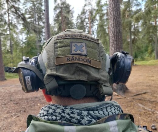Taistelijalla on päässään kuulosuojaimet ja kypärä. Kypärässä on merkkejä, joissa lukee "maakuntajoukot" ja "rändom".