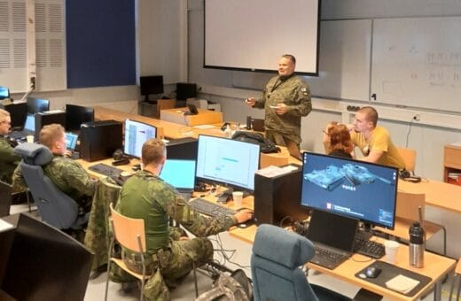 Maastoasuisia ihmisiä luokassa tietokoneiden näyttöjen ääressä. Kouluttaja luokan edessä