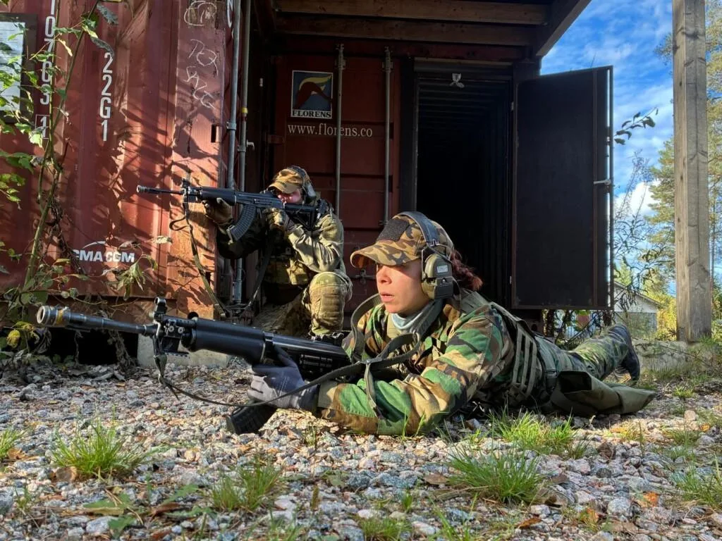 Kaksi henkilöä sotilaspuvuissa ja aseet kädessä väijyvät kontin edessä