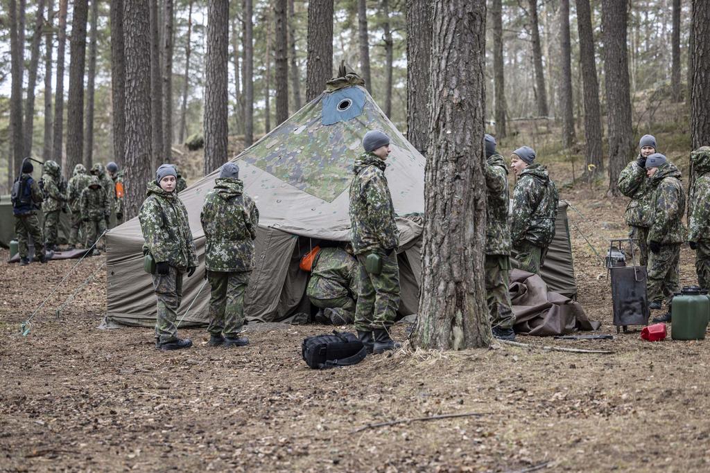 Ryhmä maastopukuisia nuoria metsässä pystyttämässä puolijoukkuetelttaa.