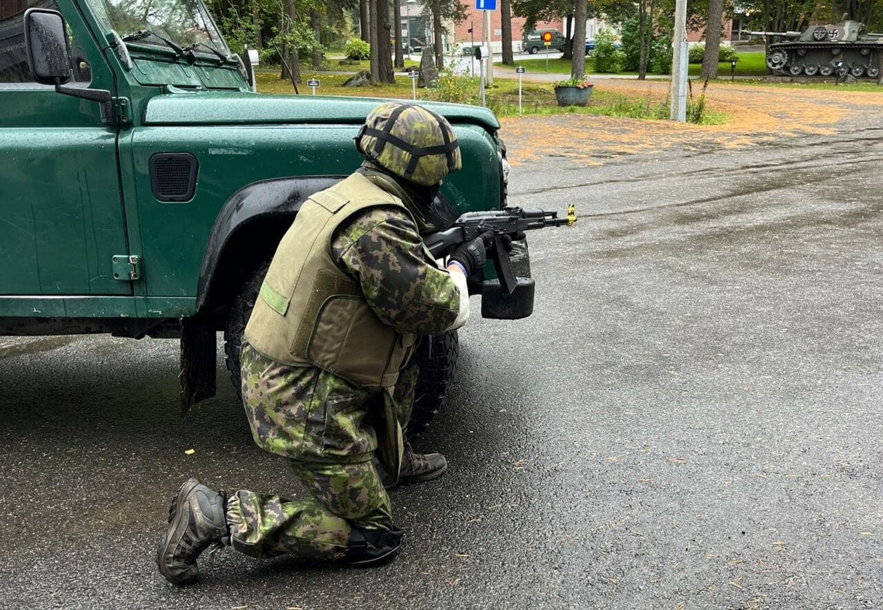 Sotilasasuinen henkilö väijyy aseen kanssa auton nokan edessä