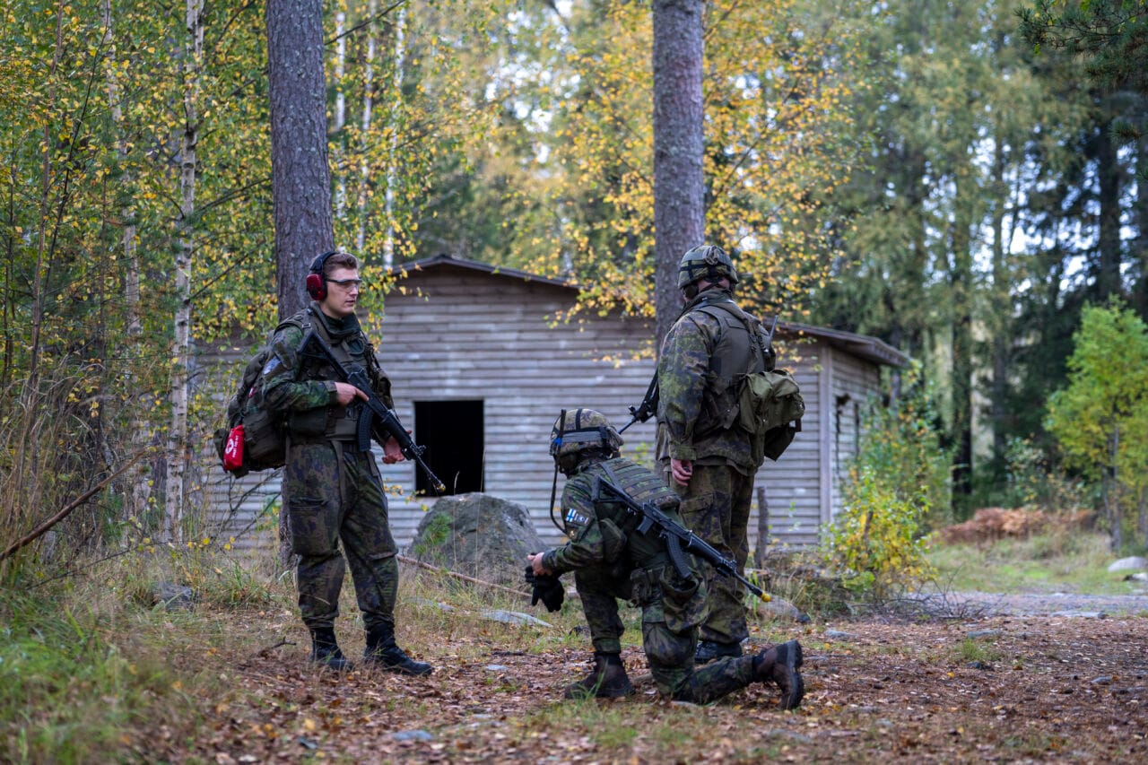 Kolme henkilöä metsässä maastopuvuissa ja aseet kädessä kohteensuojaustehtävässä.