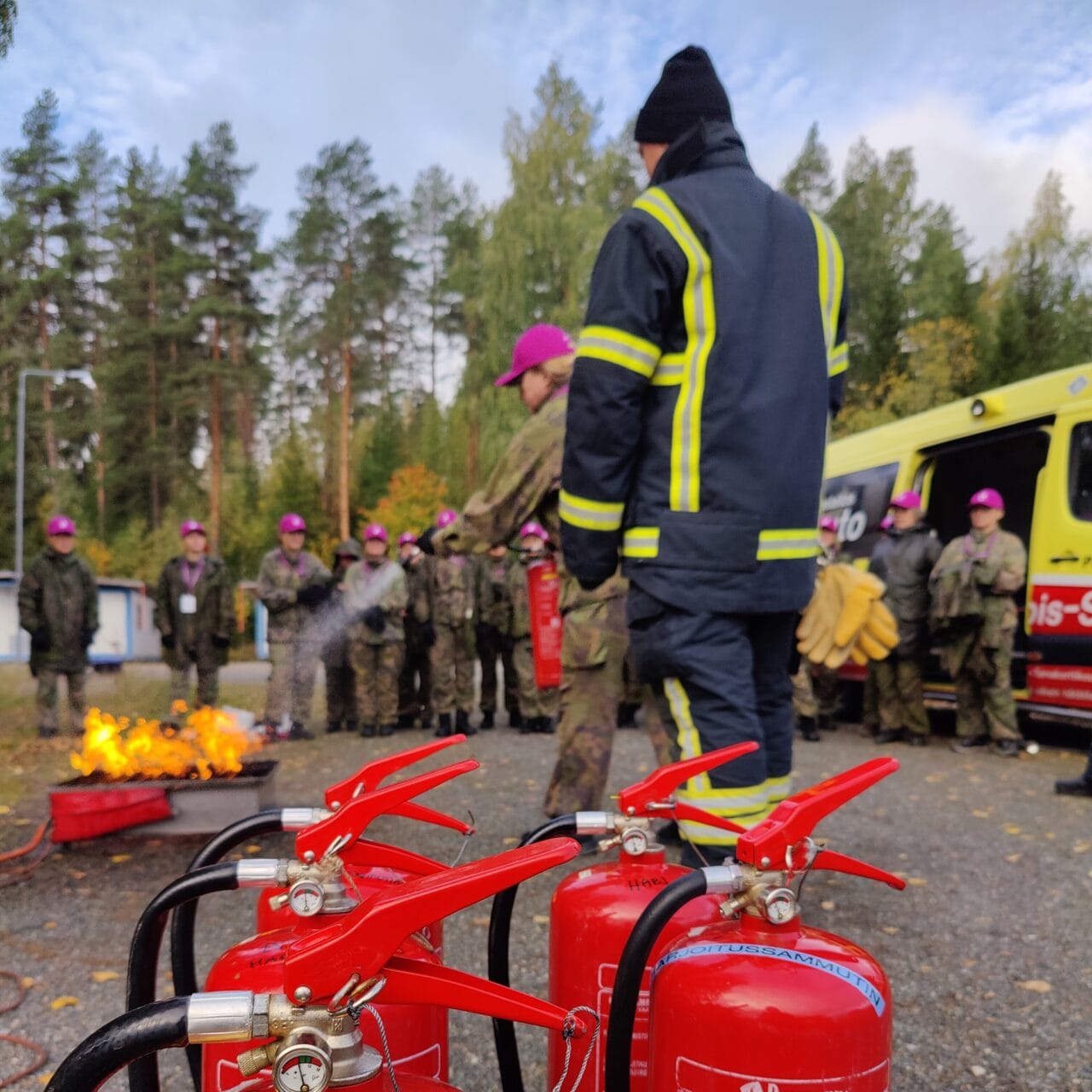Käynnissä on alkusammutuskoulutus. Etualalla neljä punaista vaahtosammutinta. Taustalla sumeana koulutettava joukko, noin parikymmentä maastoasuista naista ja heidän edessään tulipaloa sammuttava nainen. Vieressä kouluttajana pelastajan asuun pukeutunut mies.