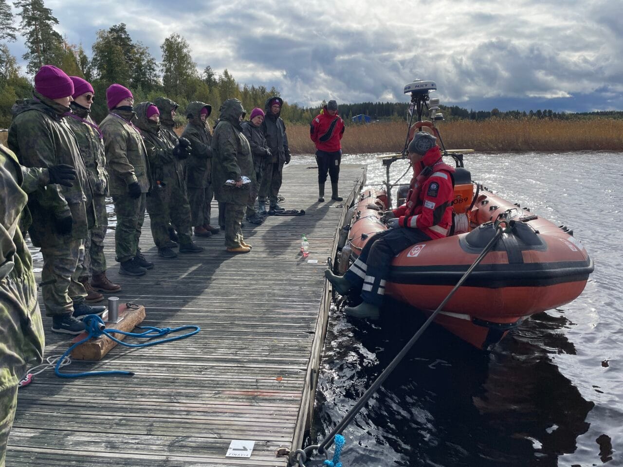 Maastopukuisia NASTA-harjoituksen osallistujia violetit pipot päässään seisomassa laiturilla. Laiturin vieressä järvipelastajien kumivene ja sen reunalla istumassa kouluttaja kelluntapuku päällään.