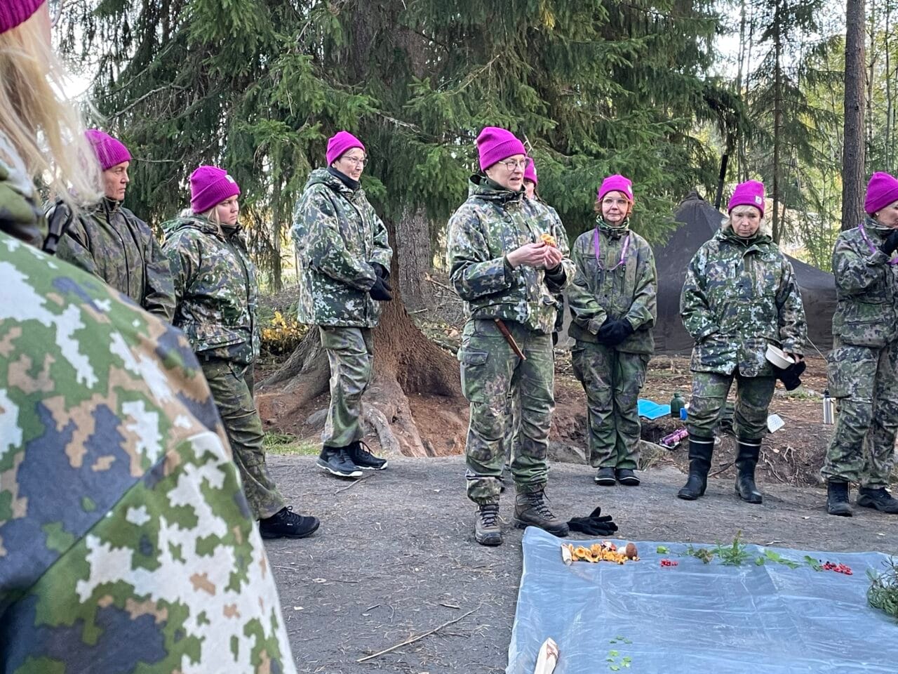 Maastopukuisia NASTA-harjoitukseen osallistujia violetit pipot päässä mäntymetsässä. Joukko puolikaaressa, keskellä kouluttaja esittelemässä syötäviä metsänantimia, kädessään kantarelli.