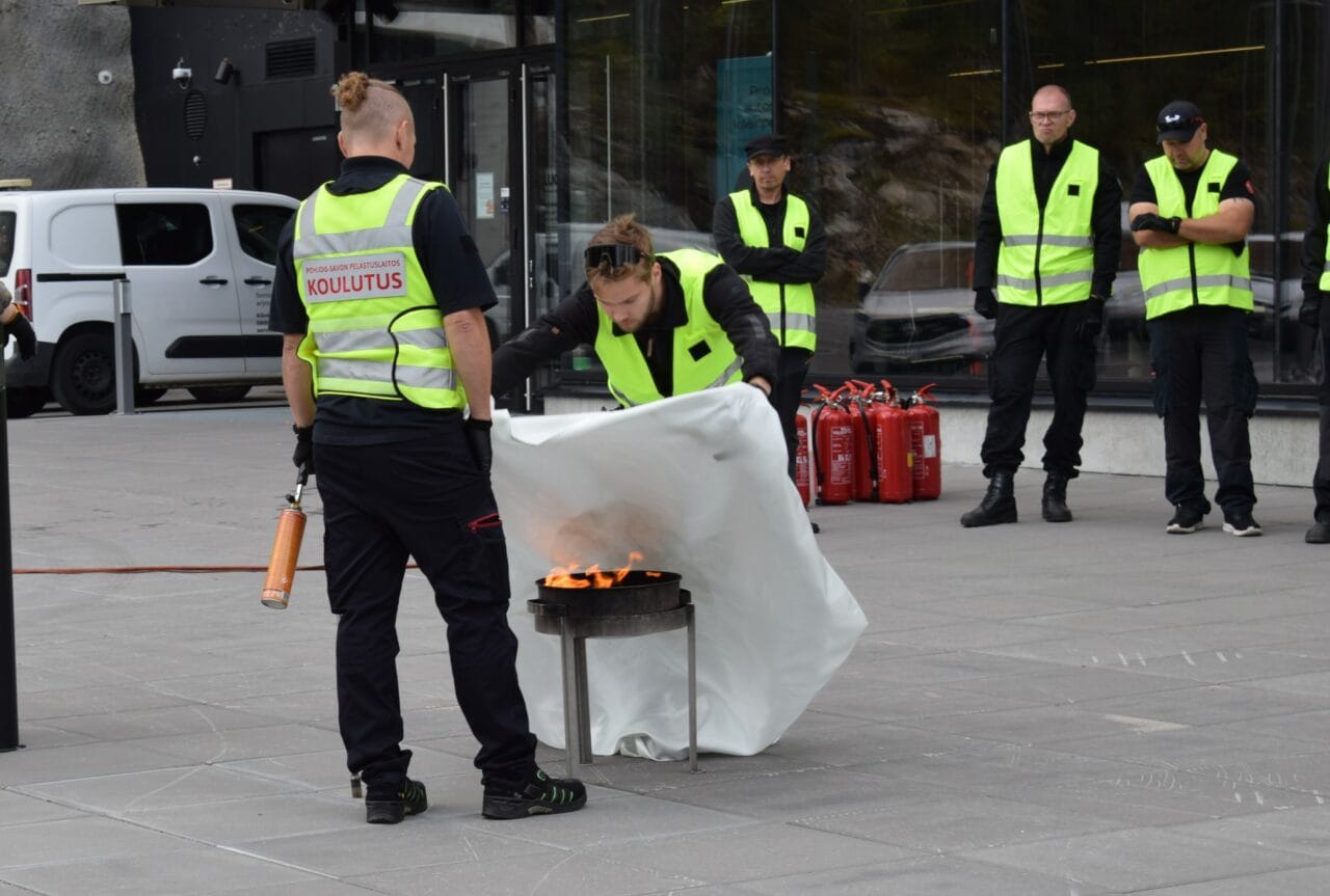 Huomioliiveihin pukeutuneet väestönsuojakouluttajat harjoittelevat alkusammutustaitoja sammutuspeitolla piha-alueella Pohjois-Savon pelastuslaitoksen kouluttajan opastuksella.