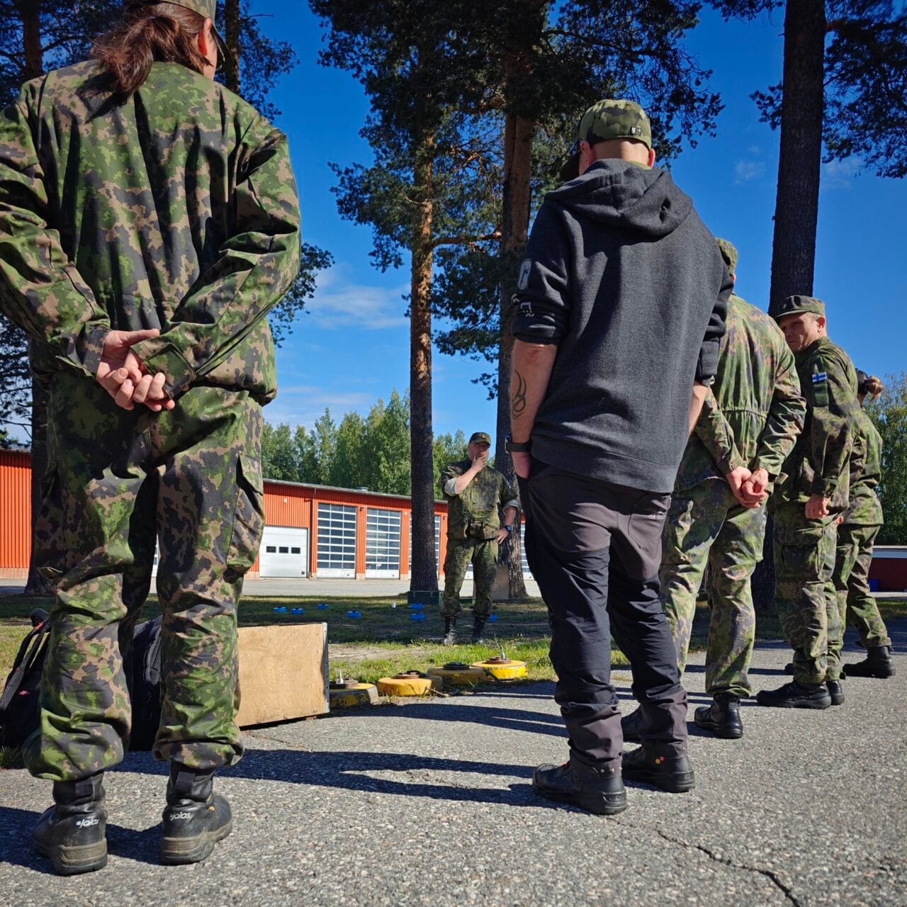 Maastopukuisia ja siviilipukuisia henkilöitä seisomassa selin kameraan rivissä ulkona aurinkoisena päivänä. Taustalla maastopukuinen kouluttaja pitämässä rastia telamiinoista.