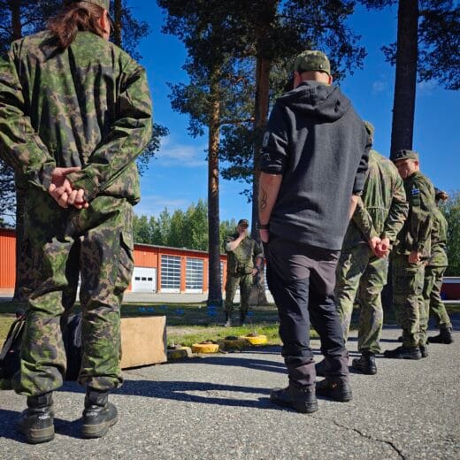 Maastopukuisia ja siviilipukuisia henkilöitä seisomassa selin kameraan rivissä ulkona aurinkoisena päivänä. Taustalla maastopukuinen kouluttaja pitämässä rastia telamiinoista.
