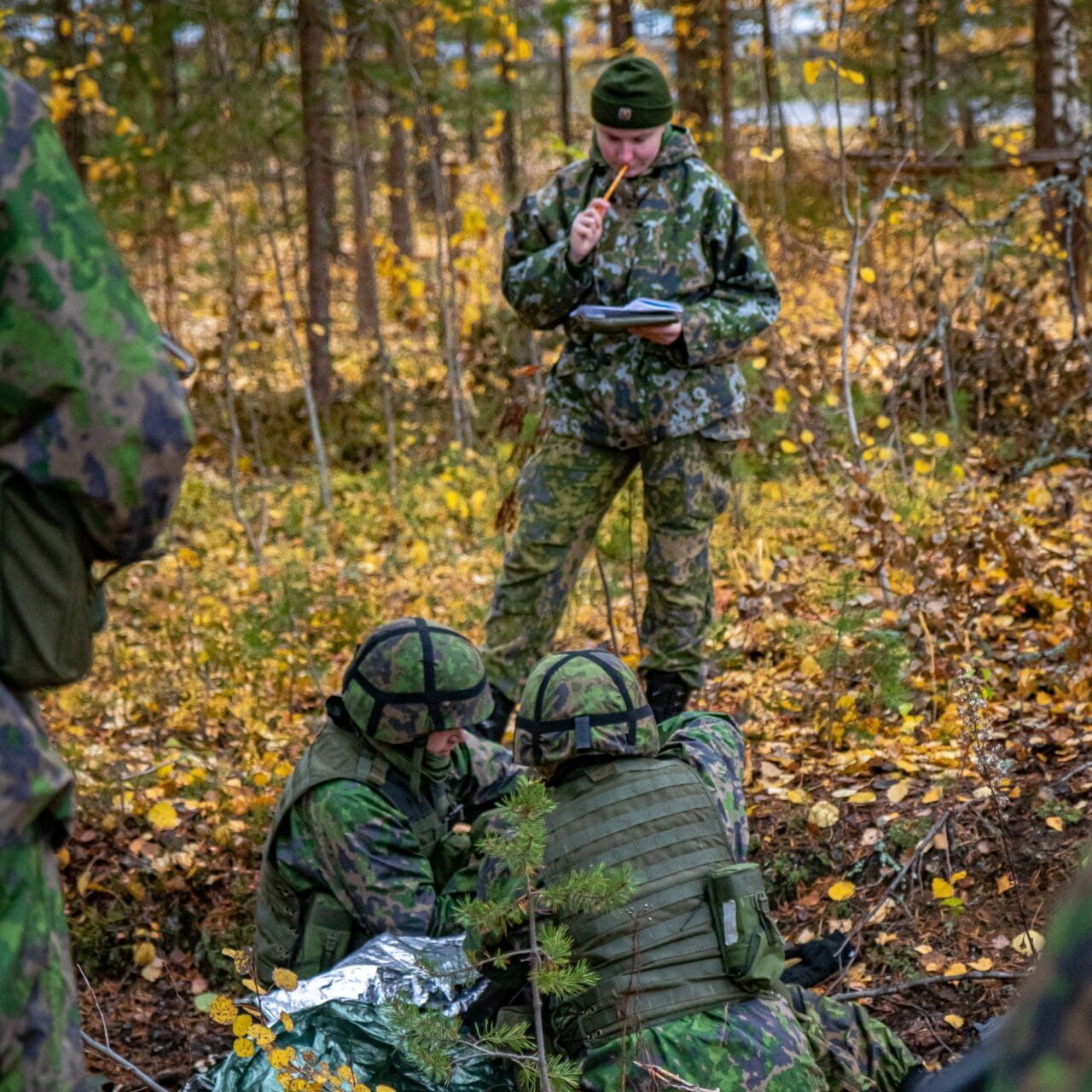 Kuvassa etualalla kaksi maastopukuista sotilasta antamassa ensiapua haavoittuneelle maakuopassa syksyisessä maastossa. Taustalla naissotilas kouluttamassa ja antamassa palautetta suoritteesta.