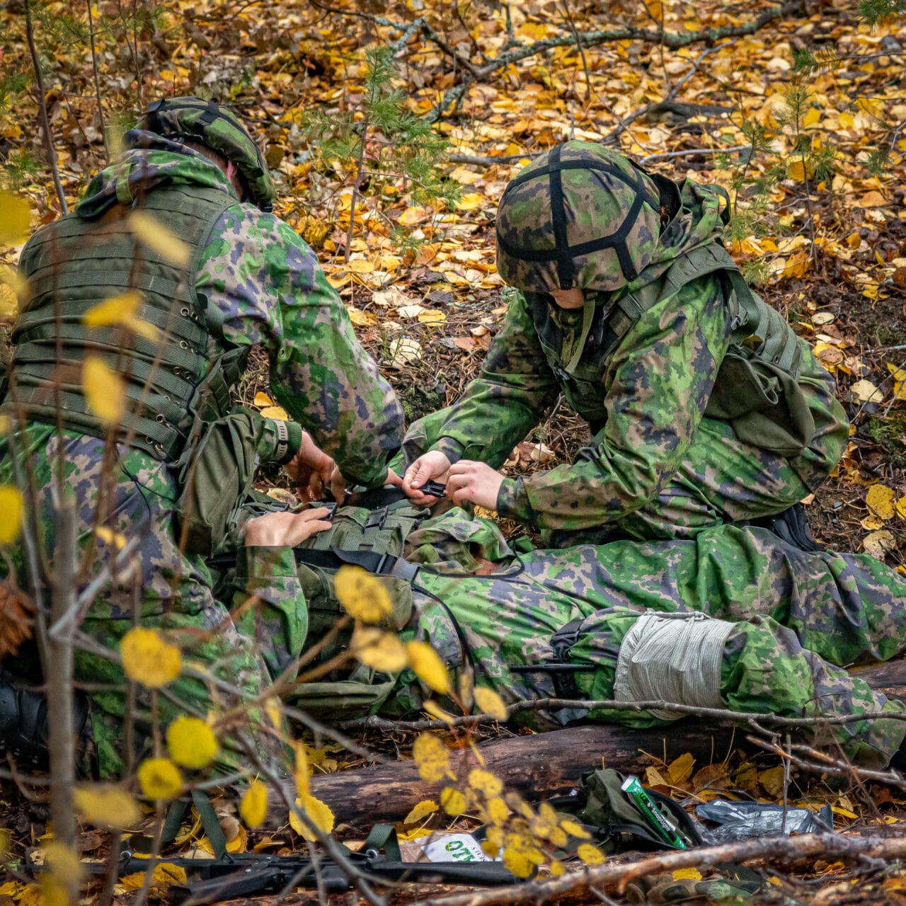 Kaksi sotilaspukuista henkilöä kenttälääkitsee kolmatta maassa makavaa sotilasta. Maasto on syksyinen