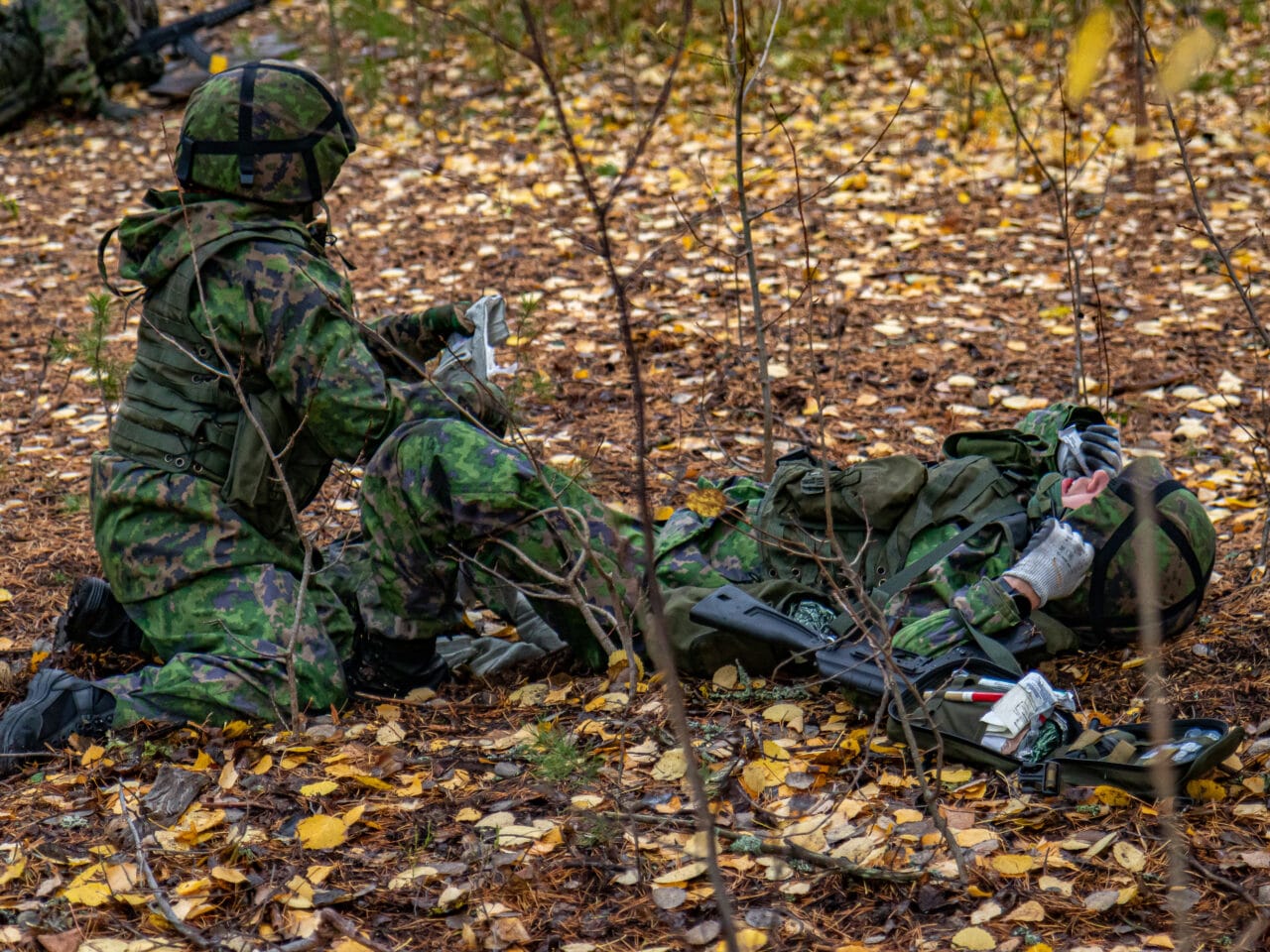 Kaksi maastopukuista sotilasta maassa syksyisessä metsässä. Yksi on haavoittunut ja makaa selällään aseensa vieressä kypärästään kiinni pitäen. Toinen sotilas on polvillaan ja on asettamassa painesidettä haavoittuneen jalkaan.