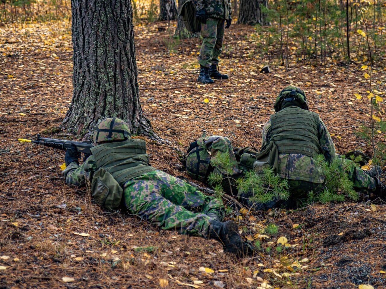 Maastopukuisia sotilaita maassa makuullaan syksyisessä maastossa. Yksi sotilaista osoittaa aseella etuviistoon maastoon ja toinen sotilas antaa haavoittuneelle ensiapua.