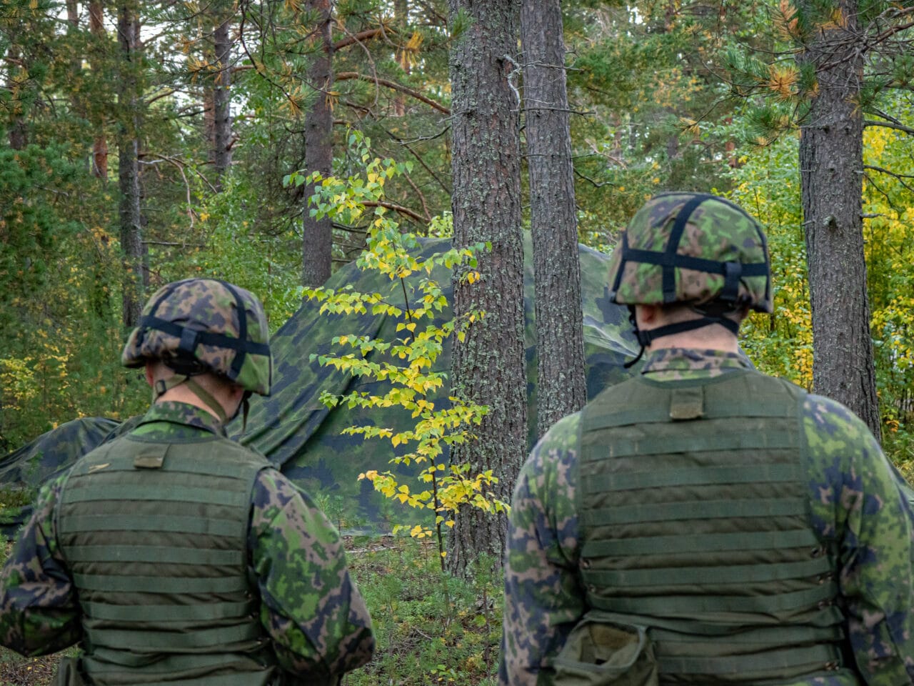Vihertävällä naamioverkolla peitettu ajoneuvo. Etualalla sumeana kaksi taisteluvarustukseen pukeutunutta sotilasta.