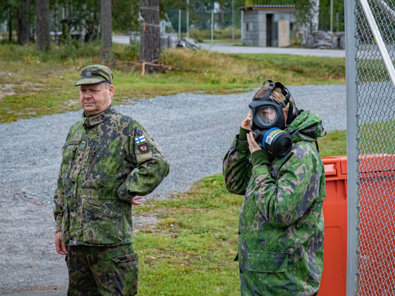 Kaksi maastopukuista sotilasta seisomassa ulkona. Etummainen on asettamassa mustaa kaasunaamaria kasvoilleen. Toinen seisoo rennommin käsi vyötäröllä.