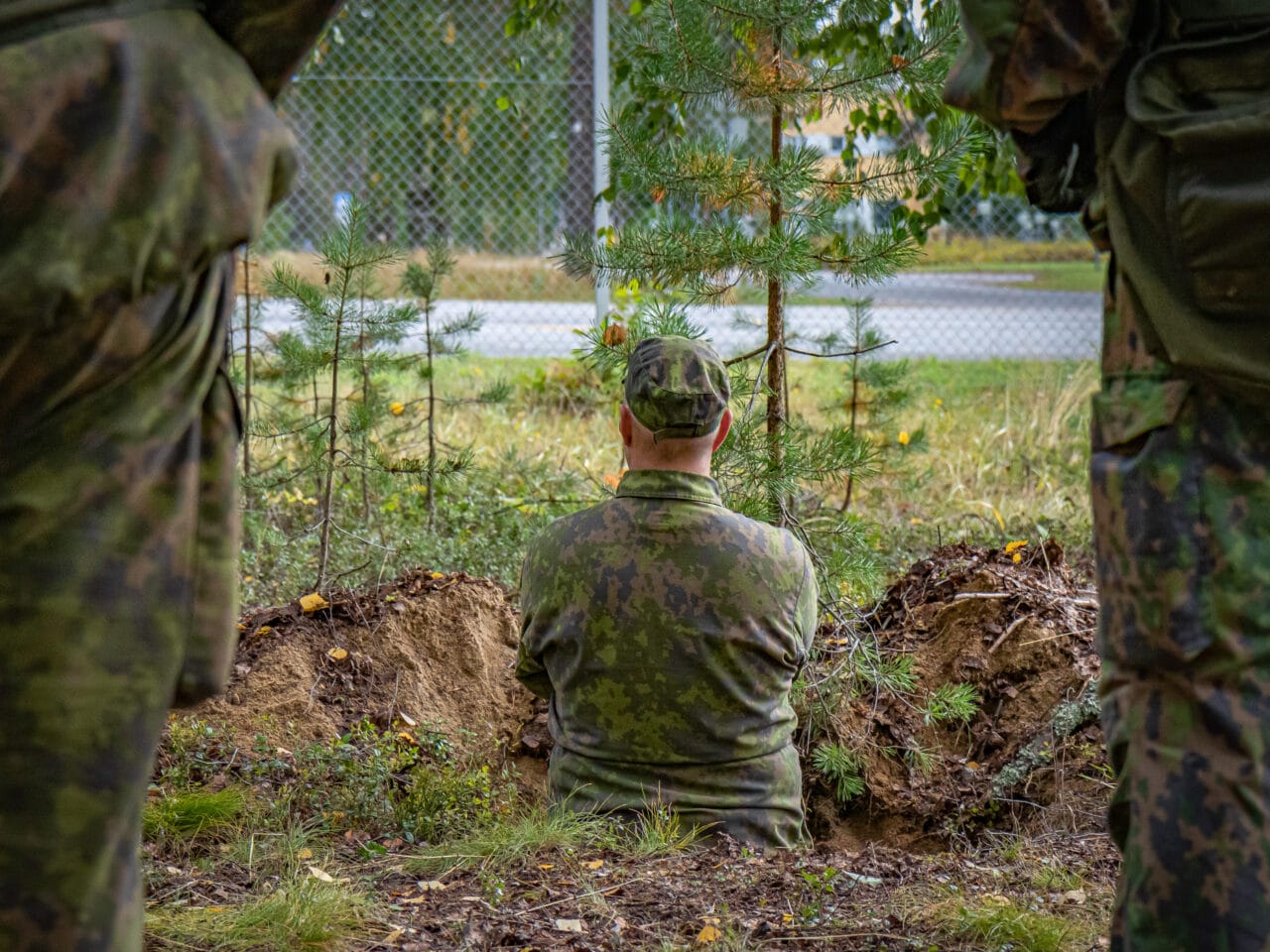 Maakuopassa seisova maastopukuinen sotilas katsomassa eteenpäin, selkä kameraan päin. Etualalla reunoilla näkyy sumeana kahden maastopukuisen ihmisen jalat.