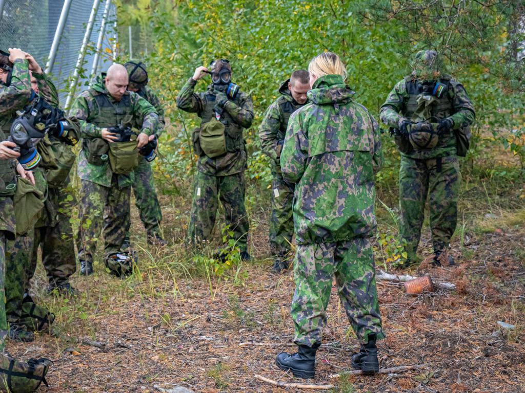 Maastopukuisia sotilaita seisomassa puoliympyrässä metsämaastossa kouluttajan edessä. Kaikilla on kädessään mustat kaasunaamarit.