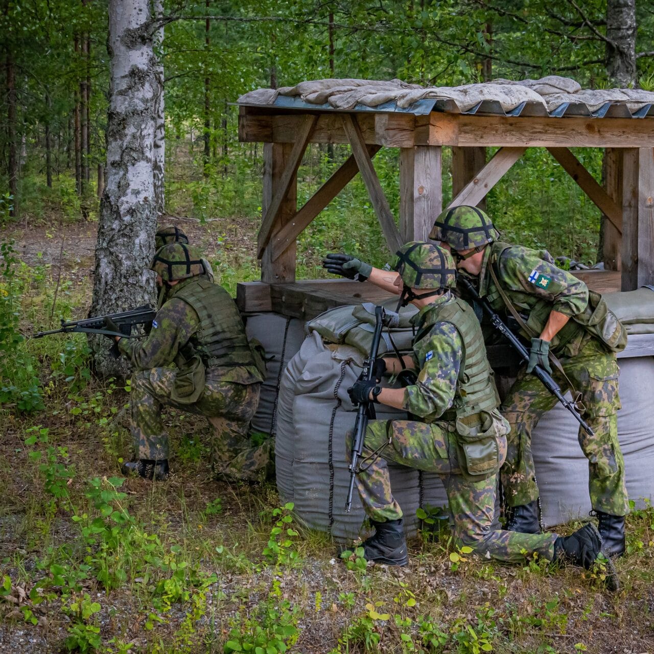 Kolme maastopukuista sotilasta polvillaan puusta ja hiekkasäkeistä rakennetun suojan vieressä. Yksi sotilaista johtaa ja osoittaa etenemissuuntaa toisille. Muut kuuntelevat.