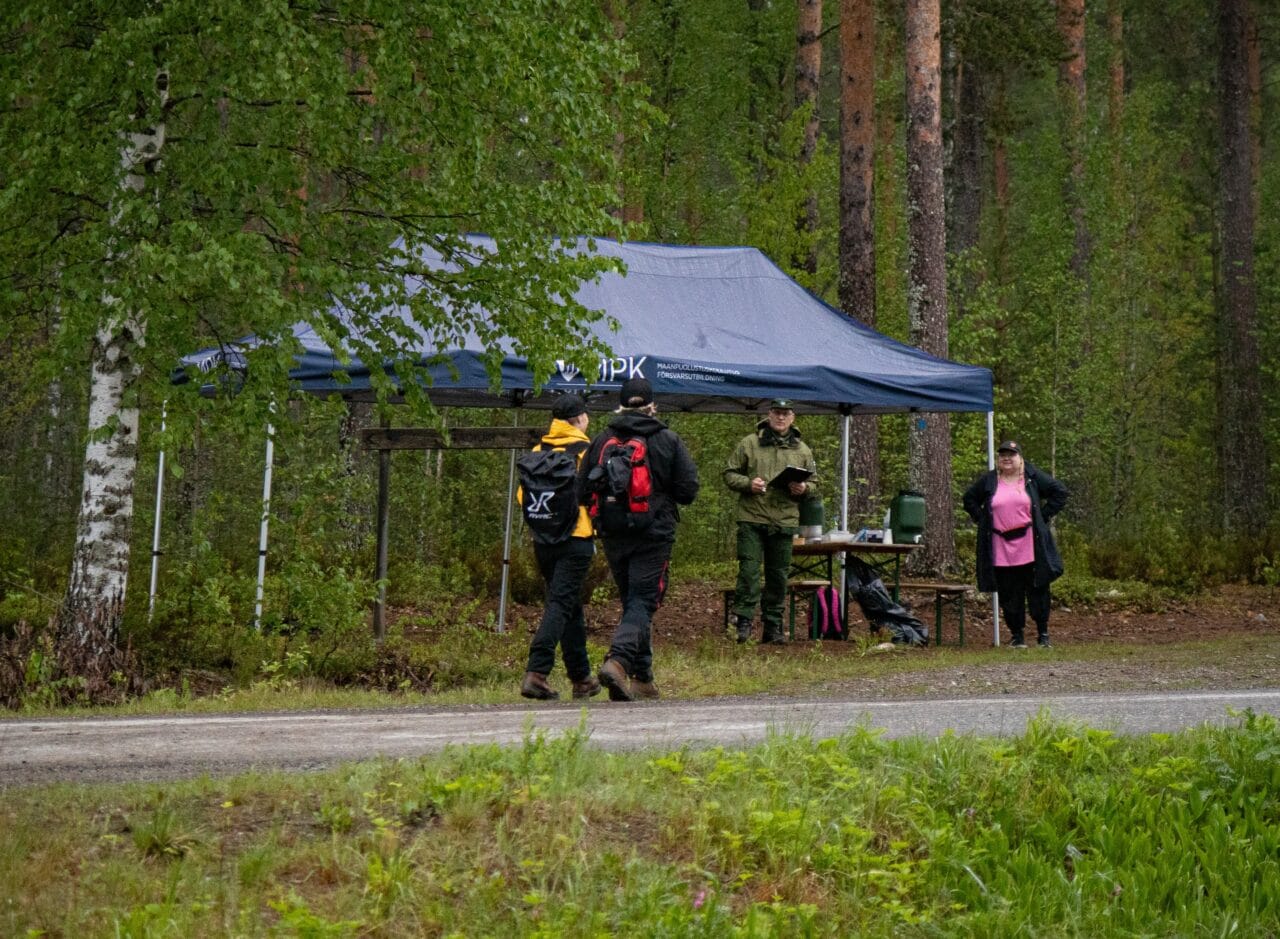 Marssijoita kävelemässä hiekkatietä pitkin mäntymetsässä. Tien vieressä taustalla MPK:n sininen teltta ja marssin huoltorasti, jossa MPK:n vihreään kenttäpukuun pukeutunut mies on nimilistan kanssa valmiina ottamaan marssijoita vastaan. Paikalla on myös pinkkiin paitaan pukeutunut nainen, joka tarjoilee marssijoille vettä.