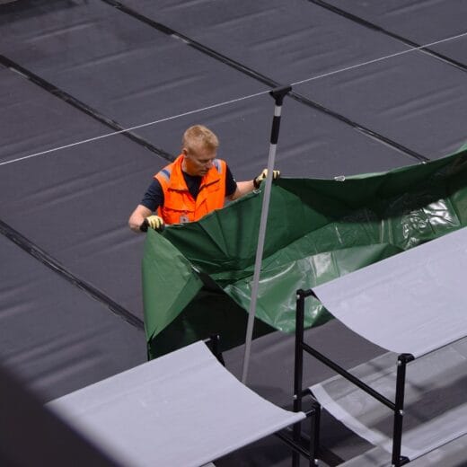 Oranssiin huomioliiviin pukeutunut mies valmistelee väestönsuojan makuuloossien väliseinän asennusta vetämällä vihreää muovipressua seinäksi.