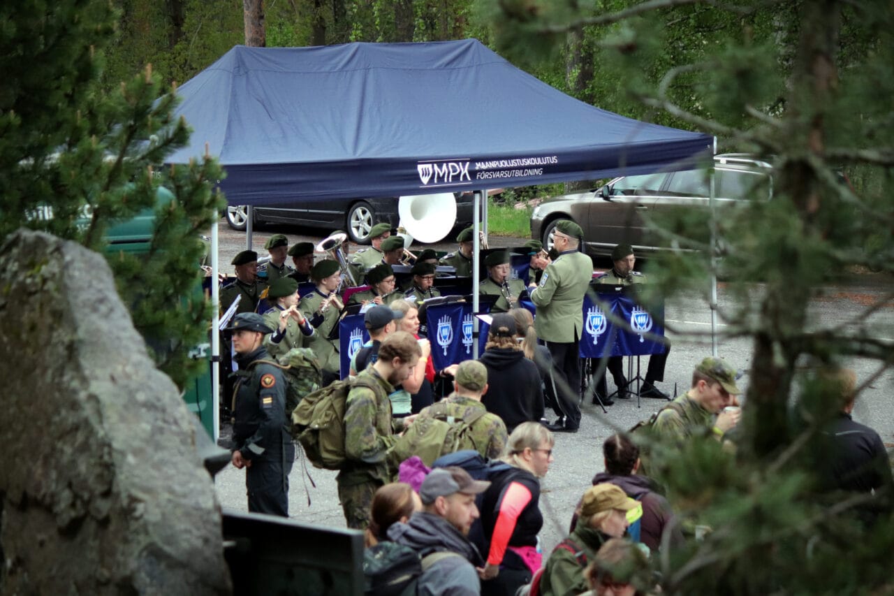 Kuvassa MPK Savo-Karjalan maanpuolustussoittokunta soittamassa sinisen teltan alla vihreissä soittoasuissaan. Etualalla sumeana marssitapahtumaan osallistuvia ihmisiä ja havupuita.